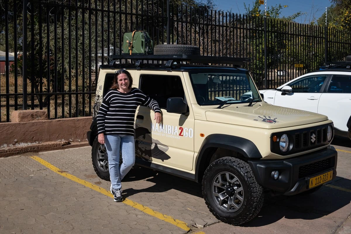 Isabelle posant devant le Suzuki Jimny 4x4 beige propre sur le parking de Namibia2go à Windhoek, avec bidon d'essence et roue secours sur le toit