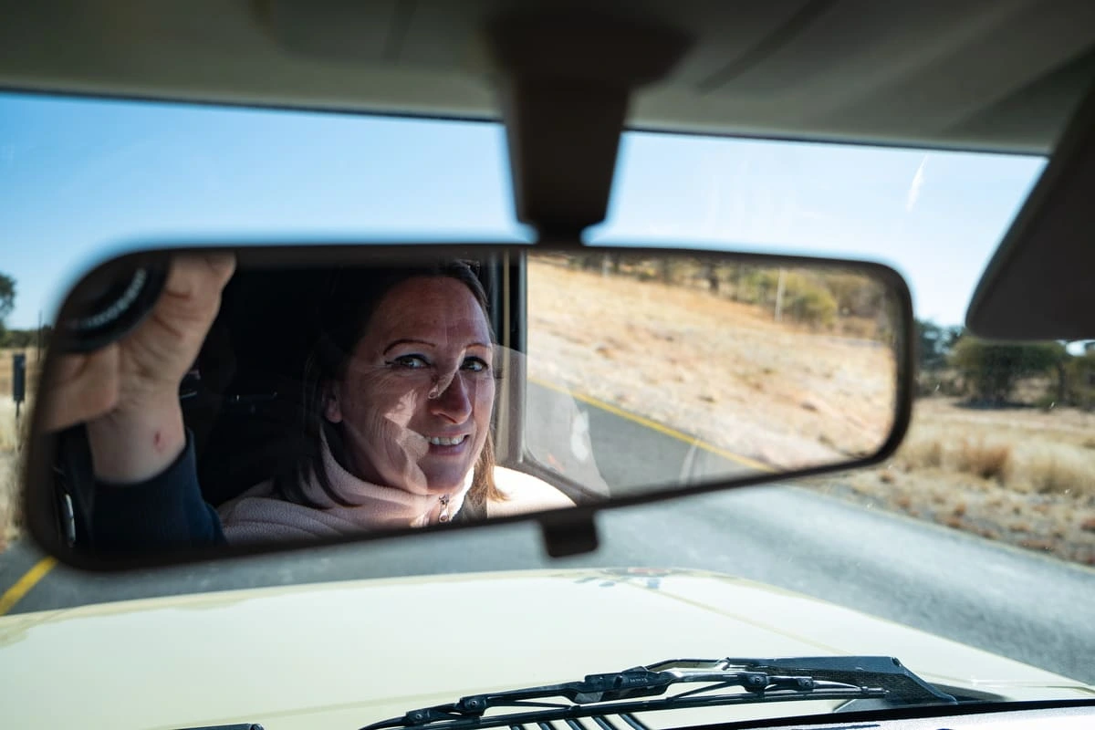 Isabelle sourit dans le rétroviseur intérieur d'un 4x4 beige sur une route en Namibie