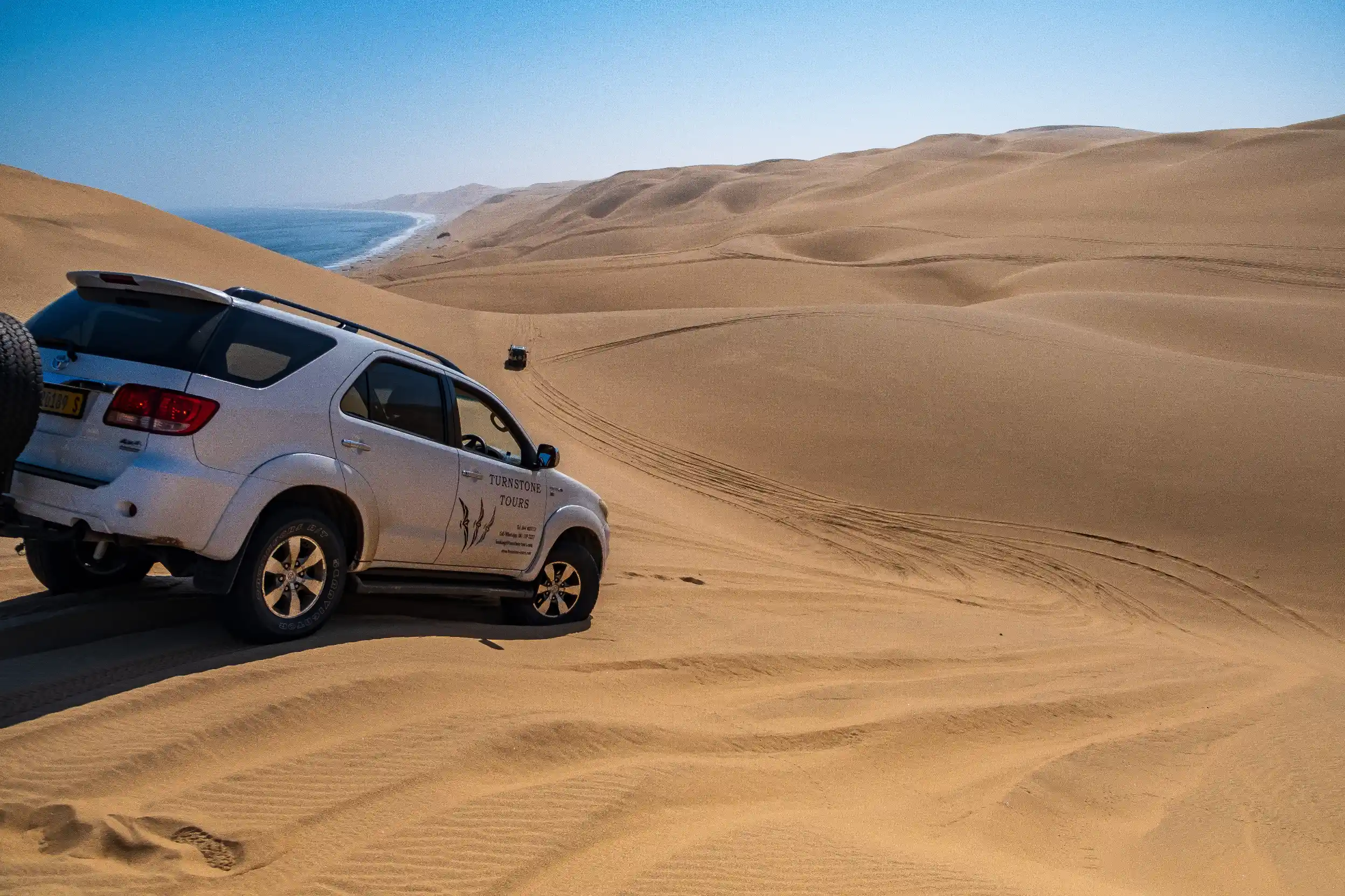 Un 4x4 blanc garé au sommet d'une dune de sable géante surplombant l'océan Atlantique à Sandwich Harbour.