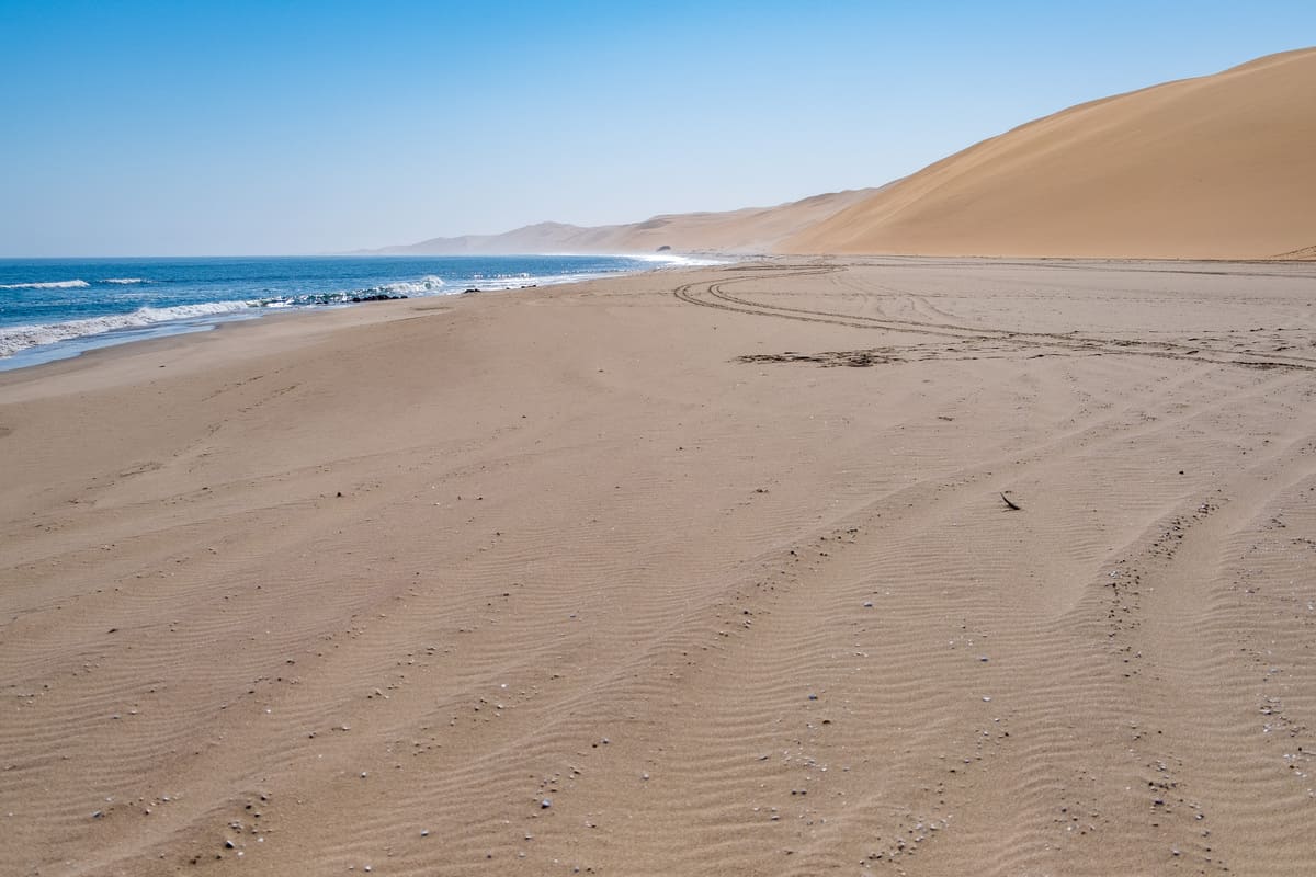 Traces de pneus de 4x4 sur une large plage de sable bordée par l'océan Atlantique et de hautes dunes à Sandwich Harbour
