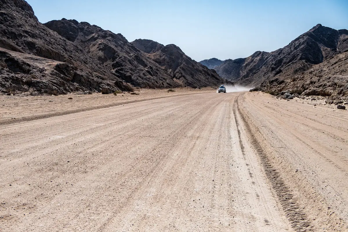 Un 4x4 soulevant la poussière sur une large piste de sable blanc traversant un canyon aride en Namibie.